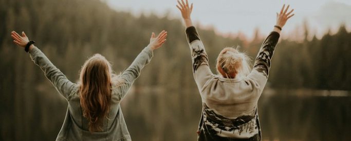 2 women with their hands up standing by a lake